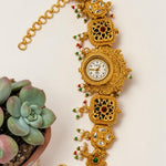 An ornate gold-toned traditional watch with intricate filigree details, small red and green bead accents, and a white dial, styled next to a small potted succulent on a light beige background.