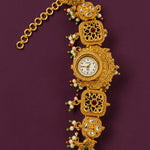 An ornate gold-toned vintage watch with a decorative link bracelet, featuring small white pearls, green and red beads, and inlaid stones, displayed on a dark purple background.