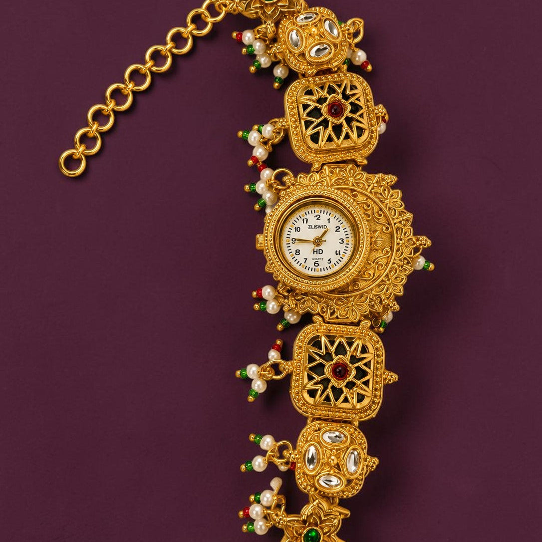 An ornate gold-toned vintage watch with a decorative link bracelet, featuring small white pearls, green and red beads, and inlaid stones, displayed on a dark purple background.