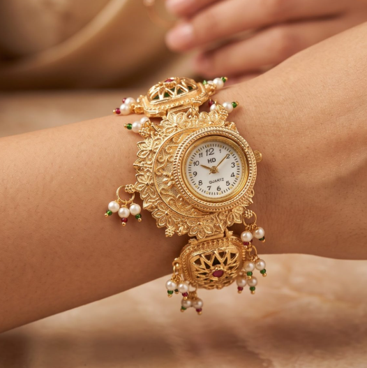 An ornate gold-plated wrist watch with intricate filigree detailing, white pearl beads, and small red and green stone accents, shown worn on a person's wrist against a warm beige background.