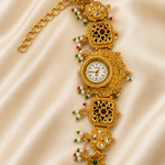 An ornate gold-toned watch bracelet with a central watch face, decorative filigree panels, small red and green stones, and pearl-like bead accents, displayed on a cream satin fabric background.