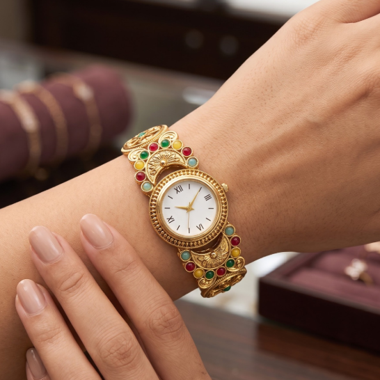 A close-up lifestyle shot of a woman's wrist wearing an ornate gold watch with a white dial and a decorative gold link bracelet embellished with red and green gemstones.