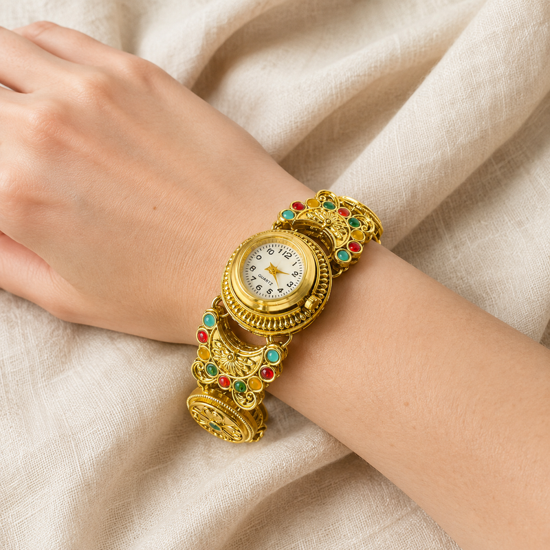 A close-up of a person's wrist wearing an ornate gold watch with a decorative link band embellished with red and green stones, set against a draped beige fabric background.