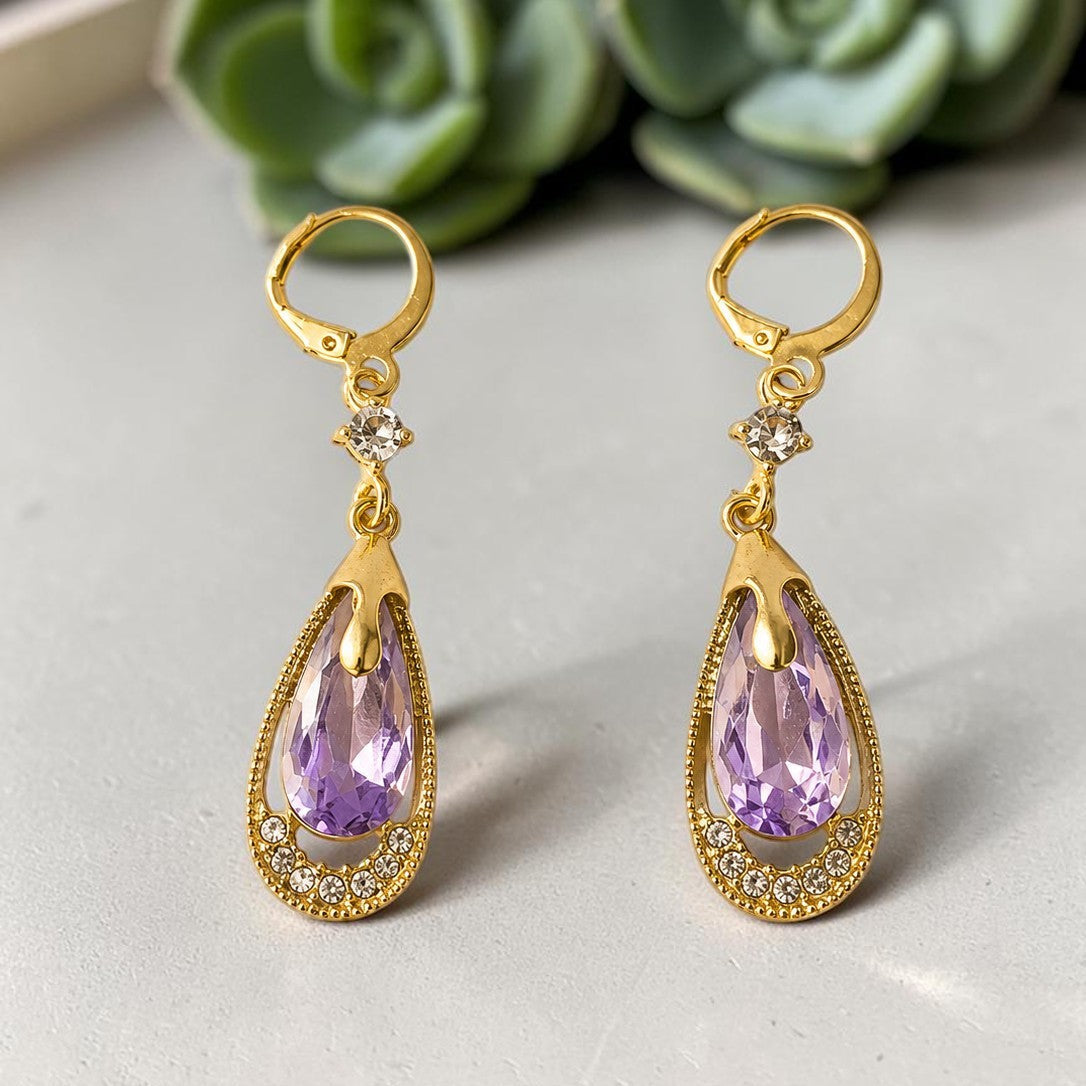 Gold tone earrings with purple teardrop-shaped gemstones on a light surface with succulents in the background.