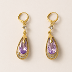 Gold tone earrings with purple teardrop-shaped gemstones on a beige background