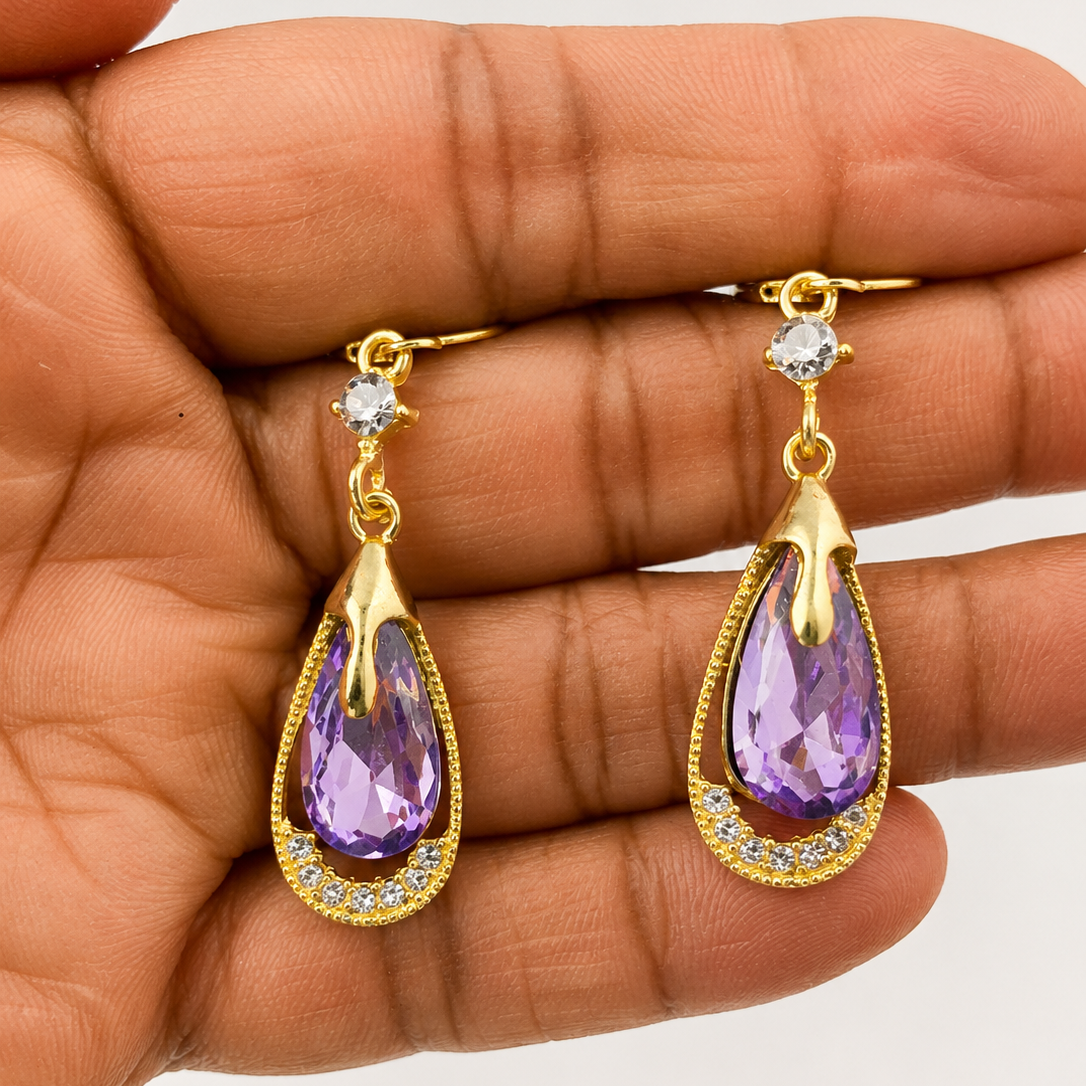 Gold tone earrings with purple teardrop stones held in a hand against a neutral background