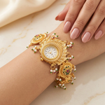 A close-up of a woman's wrist and hand wearing an ornate gold bracelet watch with a central round clock face and dangling pearl-like beads with small red and green accents, set against a soft cream satin and marble background.