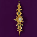An ornate gold-toned wall clock with intricate filigree details, green and red enamel accents, and small white gemstones, hanging against a solid dark purple background.