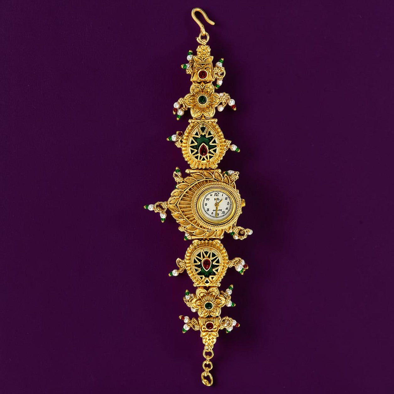 An ornate gold-toned wall clock with intricate filigree details, green and red enamel accents, and small white gemstones, hanging against a solid dark purple background.