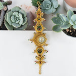 An ornate gold-plated wall clock with intricate floral engravings, small red and green gemstone accents, and a white clock face, displayed on a white surface with green succulent plants in the background.