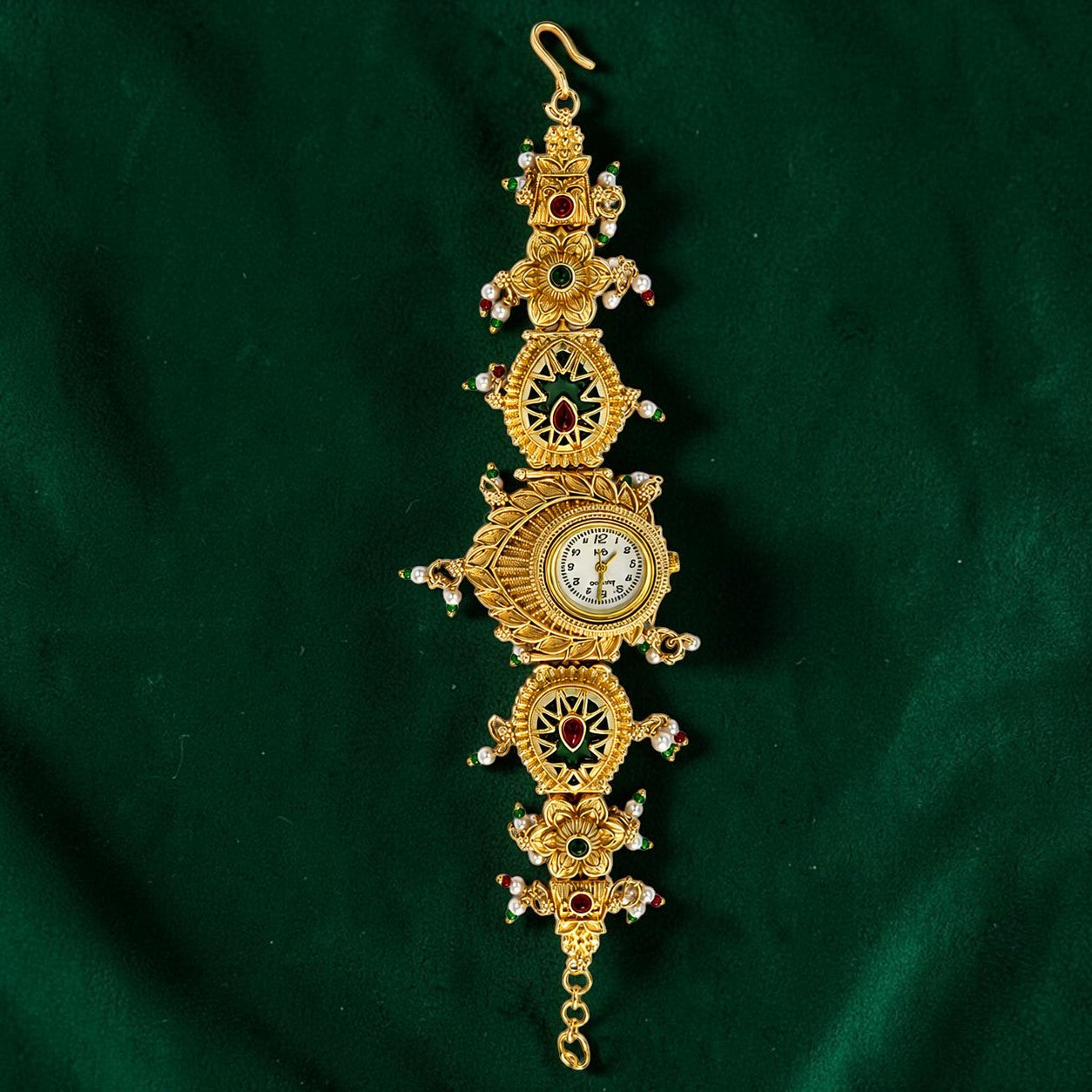 An ornate Victorian-style gold watch chain featuring a central round clock face, intricate filigree details, and small red and white gemstones, displayed on a dark green velvet background.