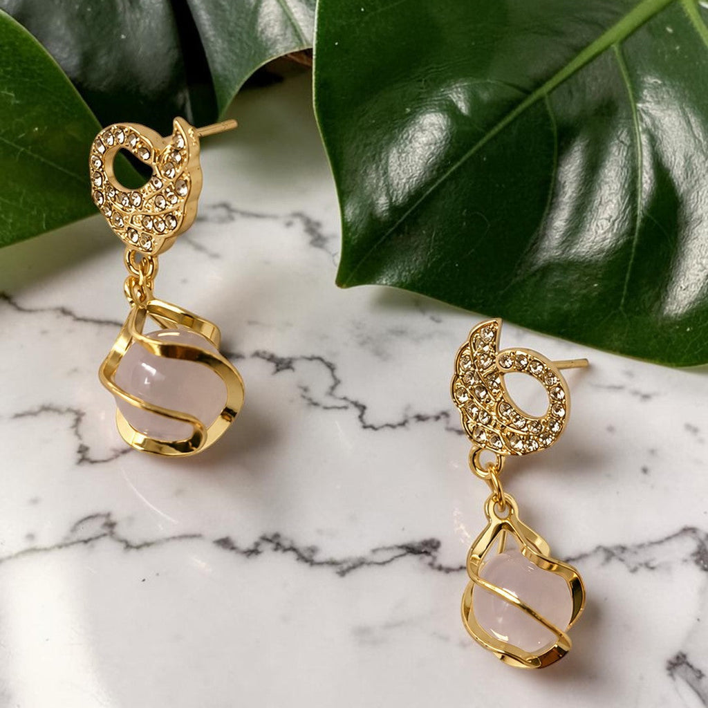 Gold tone earrings with pink stones on a marble surface with green leaves in the background