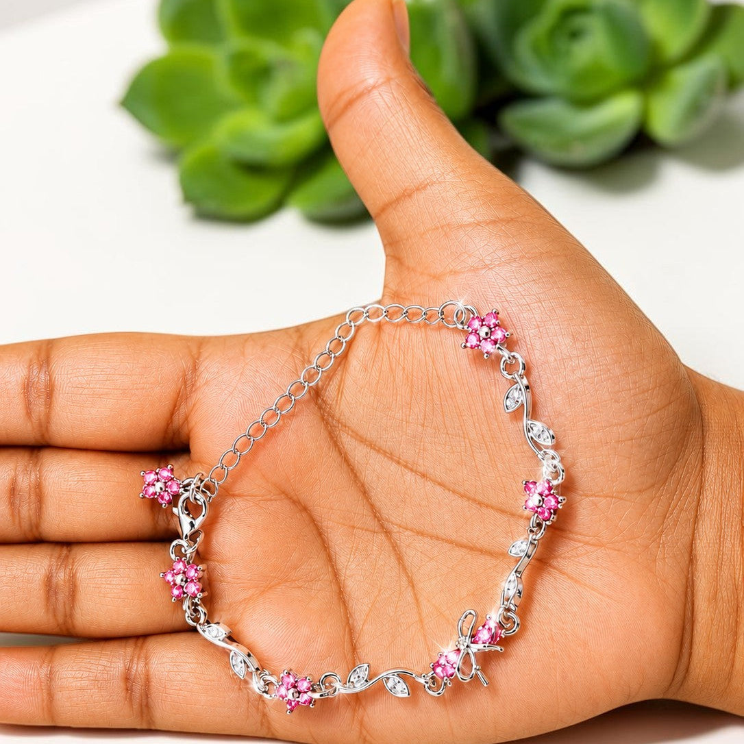 A silver chain bracelet with pink floral clusters and clear rhinestone accents, displayed on an open hand with a blurred green succulent in the background.