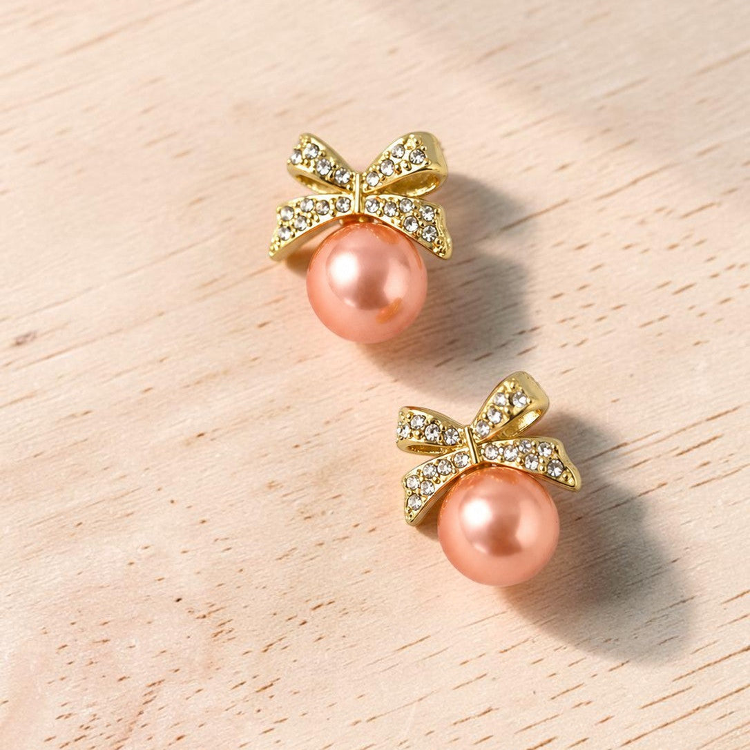 Pair of pink pearl earrings with golden bow accents on a light wooden surface