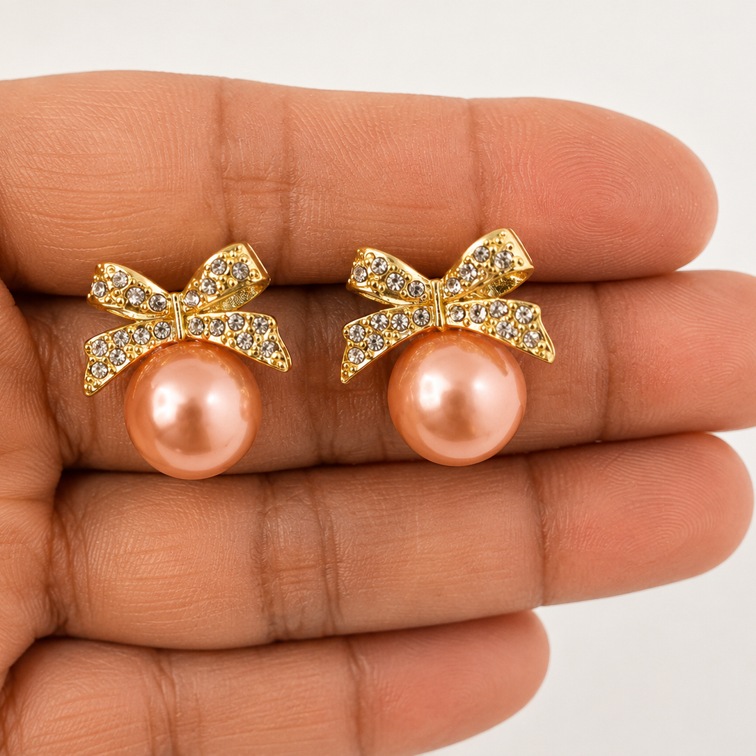 Golden bow earrings with pink pearls held between fingers on a neutral background