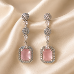 Pink square earrings with silver and crystal details on a beige fabric background