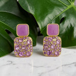 Purple square earrings with gold accents on a marble surface with green leaves in the background