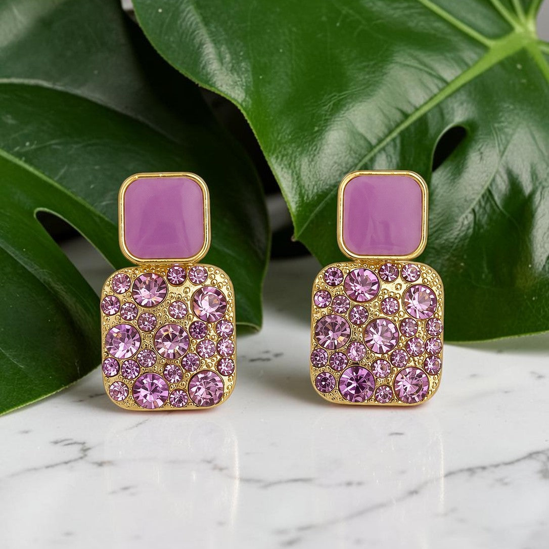 Purple square earrings with gold accents on a marble surface with green leaves in the background