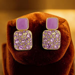 Pair of square-shaped earrings with purple stones on a brown velvet surface.