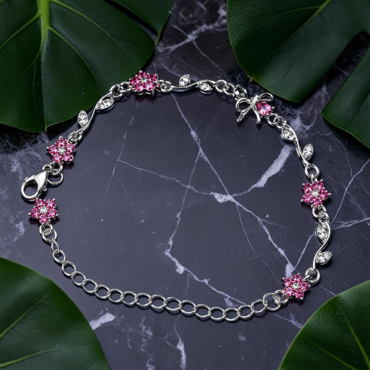 A silver chain bracelet with pink flower and bow charms, displayed on a dark marble surface surrounded by green leaves.