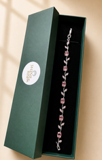 A silver-toned bracelet with pink gemstone floral motifs displayed inside an open dark green jewelry box with a white circular 'Shine Rivers' logo, set against a warm beige background with soft window shadow overlays.