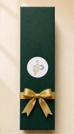 A tall, dark green rectangular gift box with a white circular label and a gold ribbon bow, photographed on a warm beige background with soft window-shadow lines.