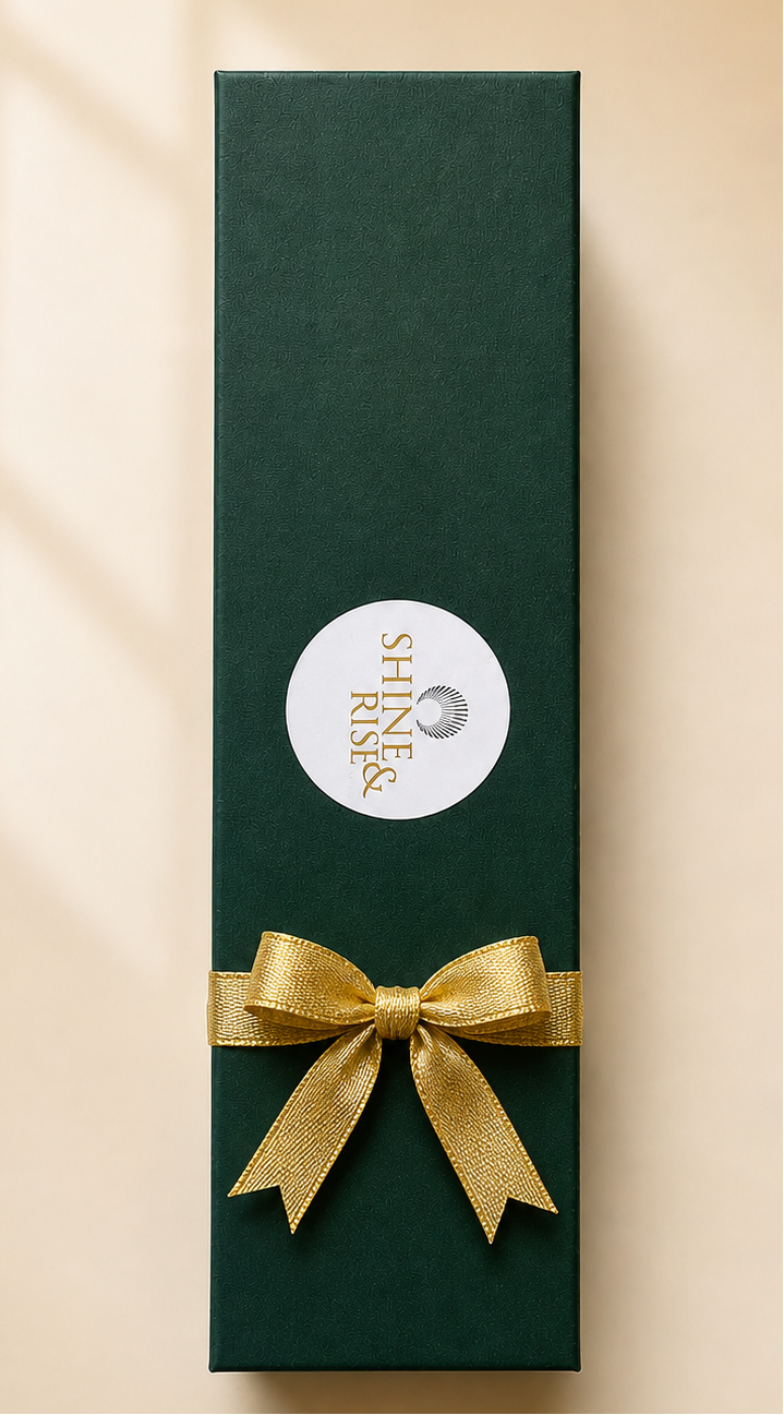 A tall, dark green rectangular gift box with a white circular label and a gold ribbon bow, photographed on a warm beige background with soft window-shadow lines.
