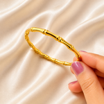 A gold bangle bracelet with a bamboo stalk design, held by a hand with pink manicured nails against a draped cream satin fabric background.