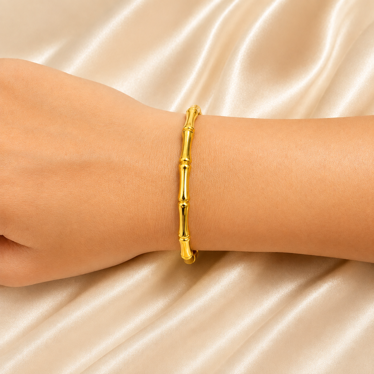 A gold bamboo bangle bracelet worn on a person's wrist, set against a draped champagne satin fabric background.