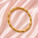 A gold-toned bangle bracelet designed to look like a bamboo stalk, featuring segmented joints, displayed on a soft pink satin fabric background.