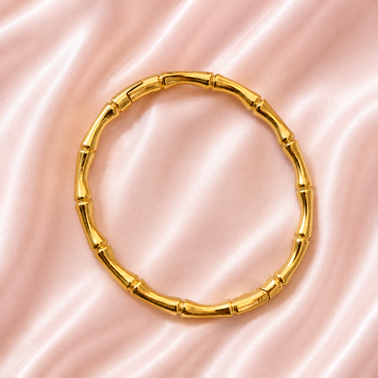 A gold-toned bangle bracelet designed to look like a bamboo stalk, featuring segmented joints, displayed on a soft pink satin fabric background.
