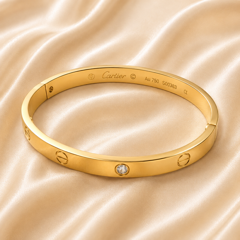 A gold bangle bracelet with screw-head details and a central clear stone, branded Cartier, resting on a draped beige satin fabric.