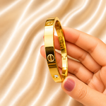 A gold bangle bracelet with screw and circle motifs, held in a hand against a draped champagne satin background.
