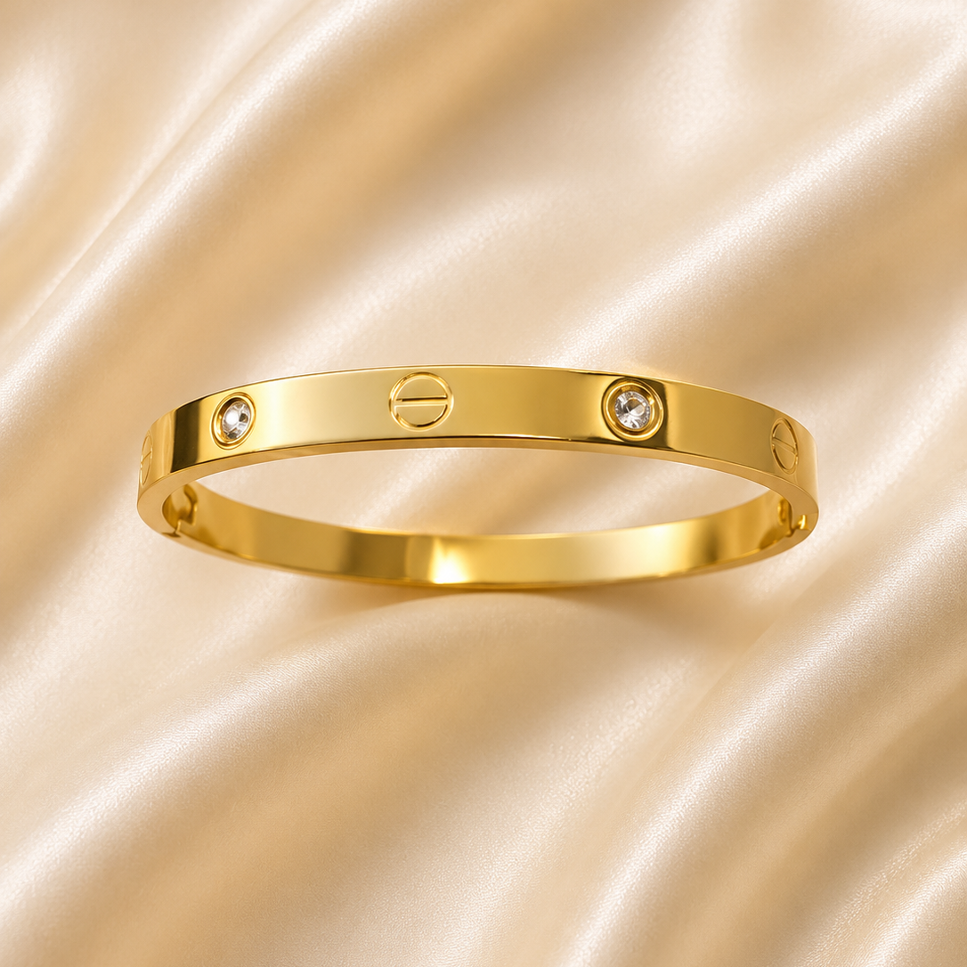 A gold bangle bracelet with two small clear stones and a screw-head motif, displayed on a draped beige satin fabric background.