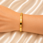 A gold bangle bracelet with three small clear gemstones, worn on a person's wrist against a draped champagne satin background.