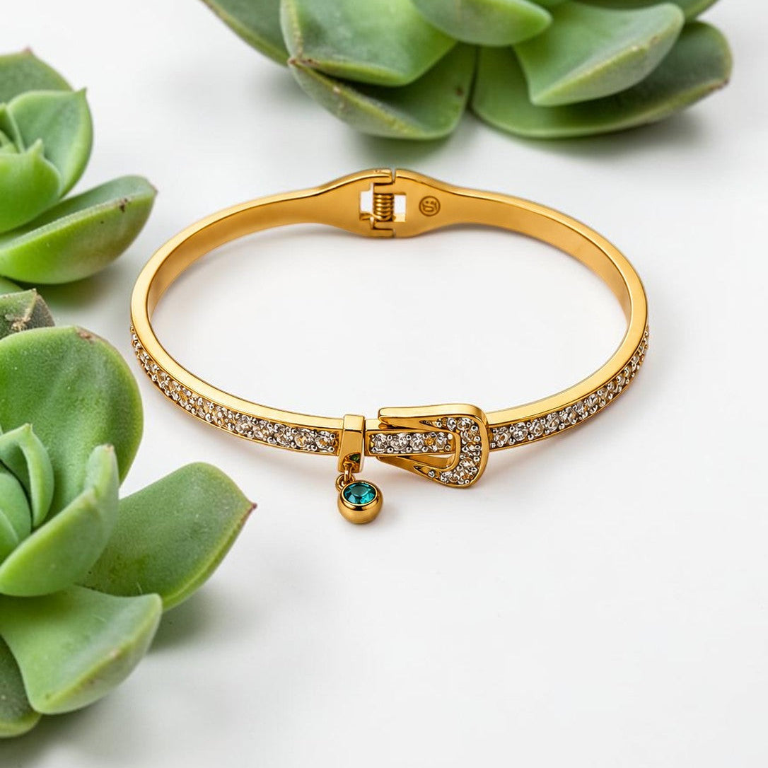 A gold-plated bangle bracelet with pavé-set clear stones and a small dangling green gemstone charm, styled with green succulent plants on a white background.