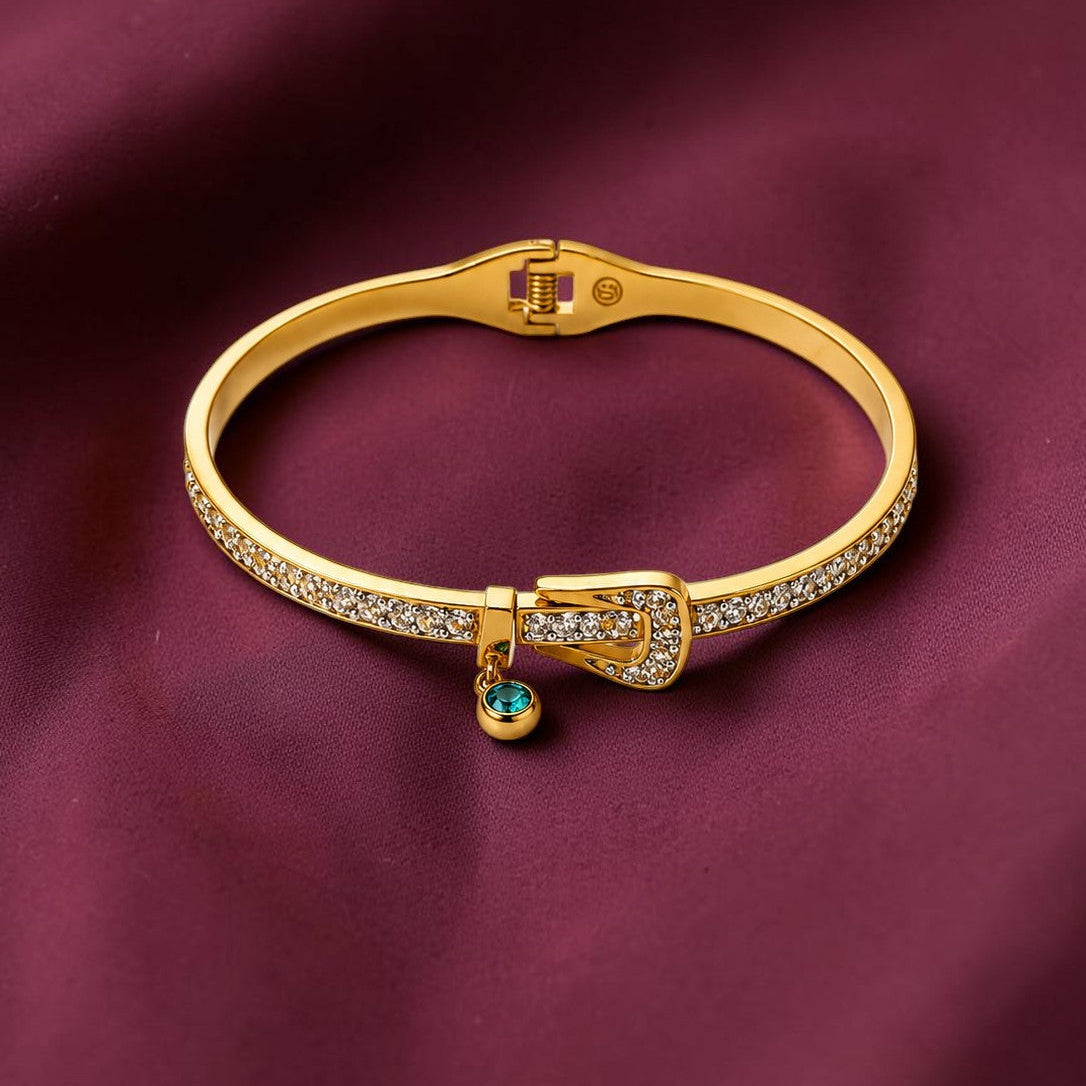 A gold-plated hinged bangle bracelet with pavé-set clear stones and a small dangling turquoise-blue bead, displayed on a draped burgundy satin fabric background.