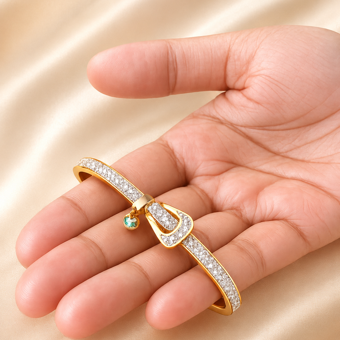A gold-toned bangle bracelet with pavé-set clear stones and a small dangling green gemstone, displayed on an open hand against a soft beige satin background.