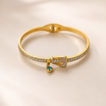 A gold bangle bracelet with pavé-set clear stones and a small dangling emerald-cut gemstone, displayed on a soft beige background.
