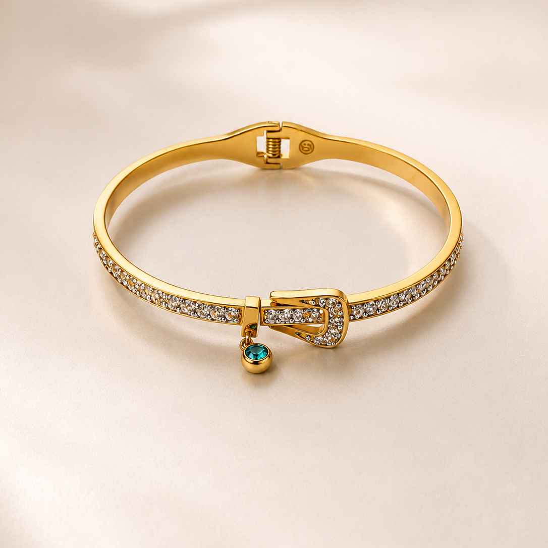 A gold bangle bracelet with pavé-set clear stones and a small dangling emerald-cut gemstone, displayed on a soft beige background.