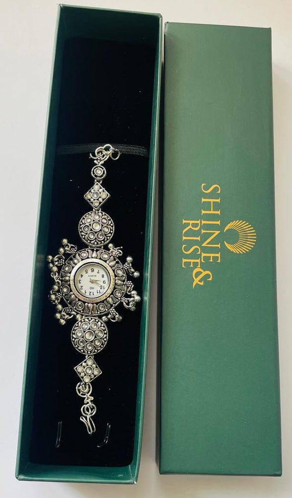 An ornate silver-toned watch with a round face and decorative crystal-like detailing, presented inside an open dark green box next to its matching green lid, which features the brand name 'SHINE & RISE' in gold.
