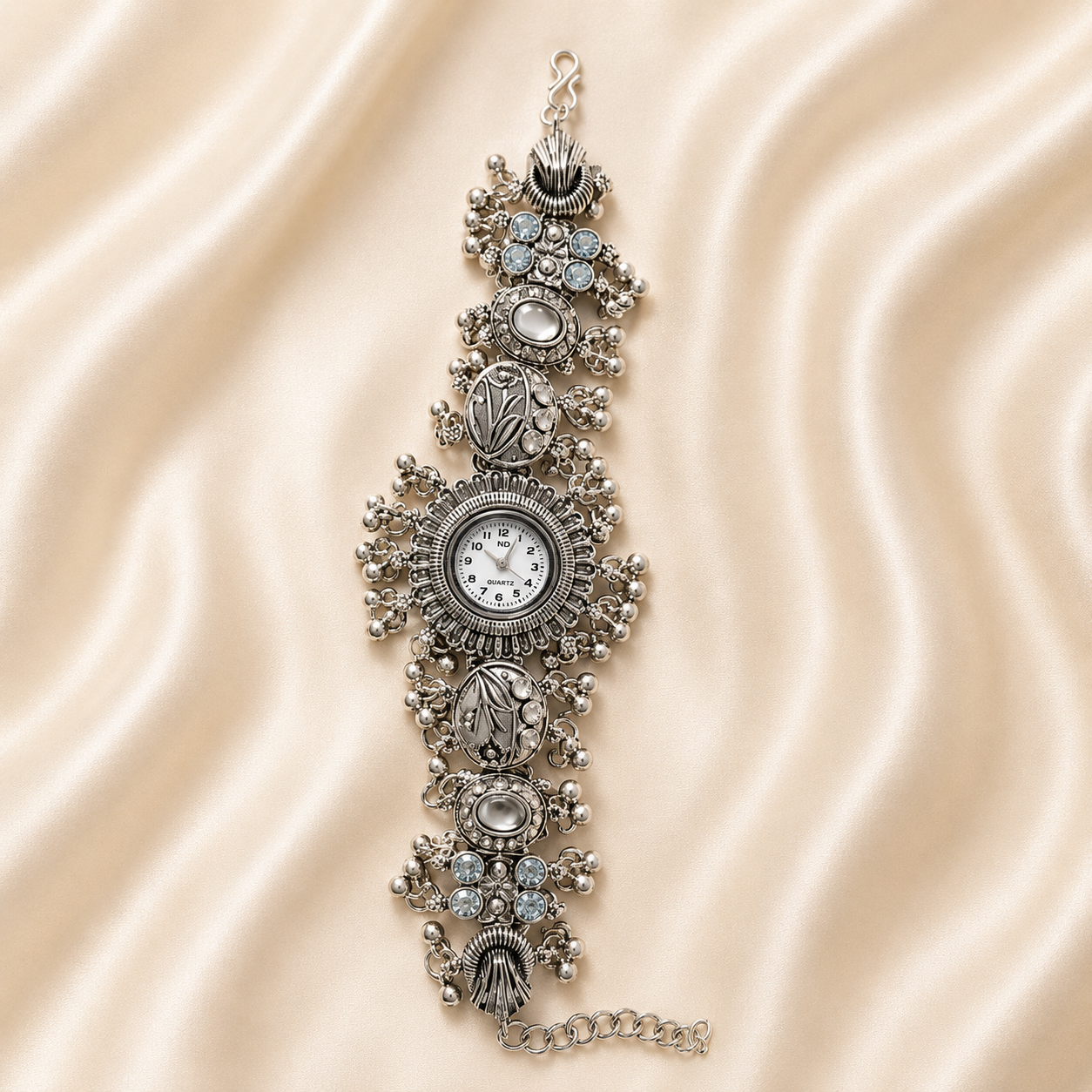 An ornate silver-toned watch bracelet featuring a central round clock face, surrounded by intricate filigree, floral motifs, and clear rhinestone embellishments, displayed on a draped beige satin fabric background.