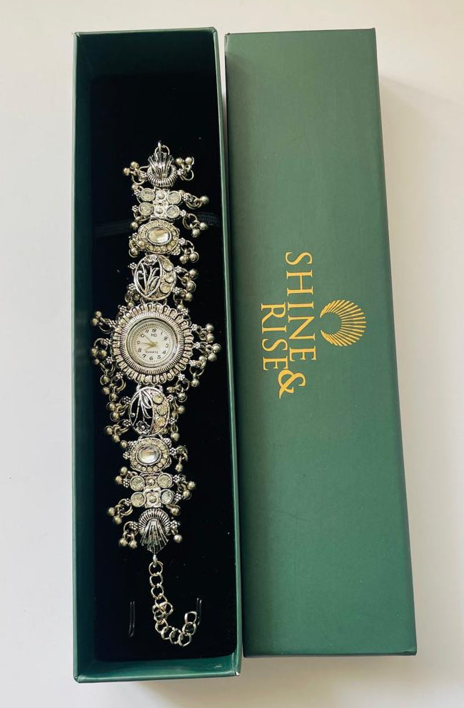 An ornate silver-toned watch with crystal embellishments, presented in an open dark green box next to its closed green lid which features the brand name 'SHINE & RISE'.
