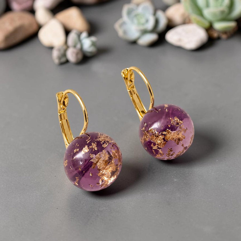 Purple earrings with gold accents on a gray surface with small plants in the background