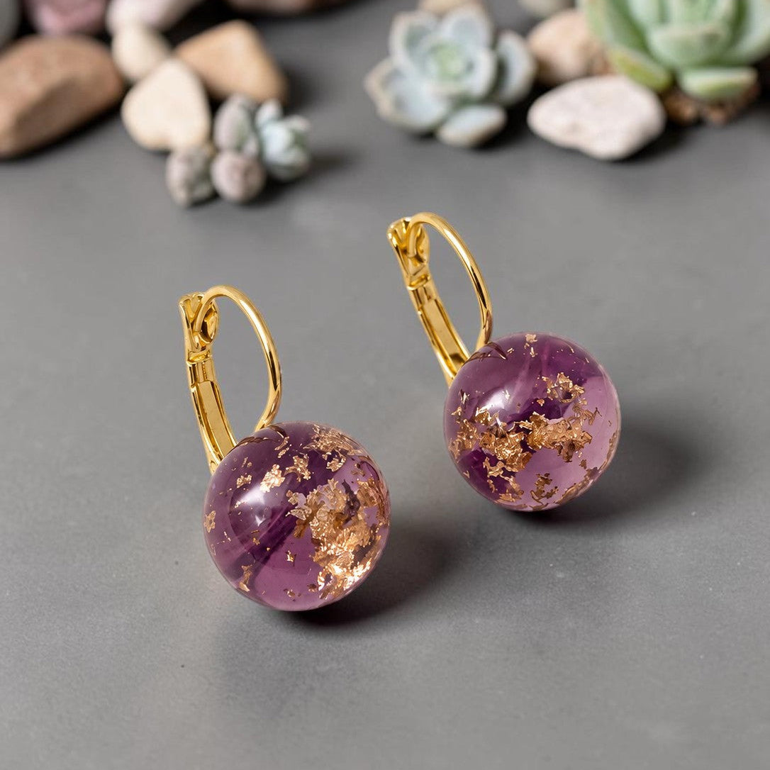 Purple earrings with gold accents on a gray surface with small plants in the background