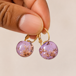 Purple earrings with gold accents held by a hand against a beige background