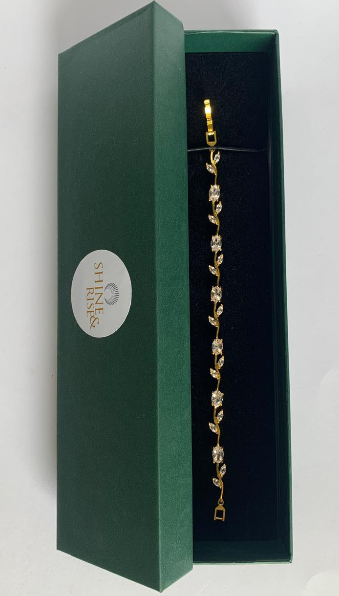 A gold leaf-patterned bracelet with clear rhinestones displayed inside an open dark green presentation box with a black velvet interior.