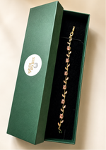 A gold bracelet with pink gemstone floral details displayed inside an open dark green jewelry box with a white circular logo.