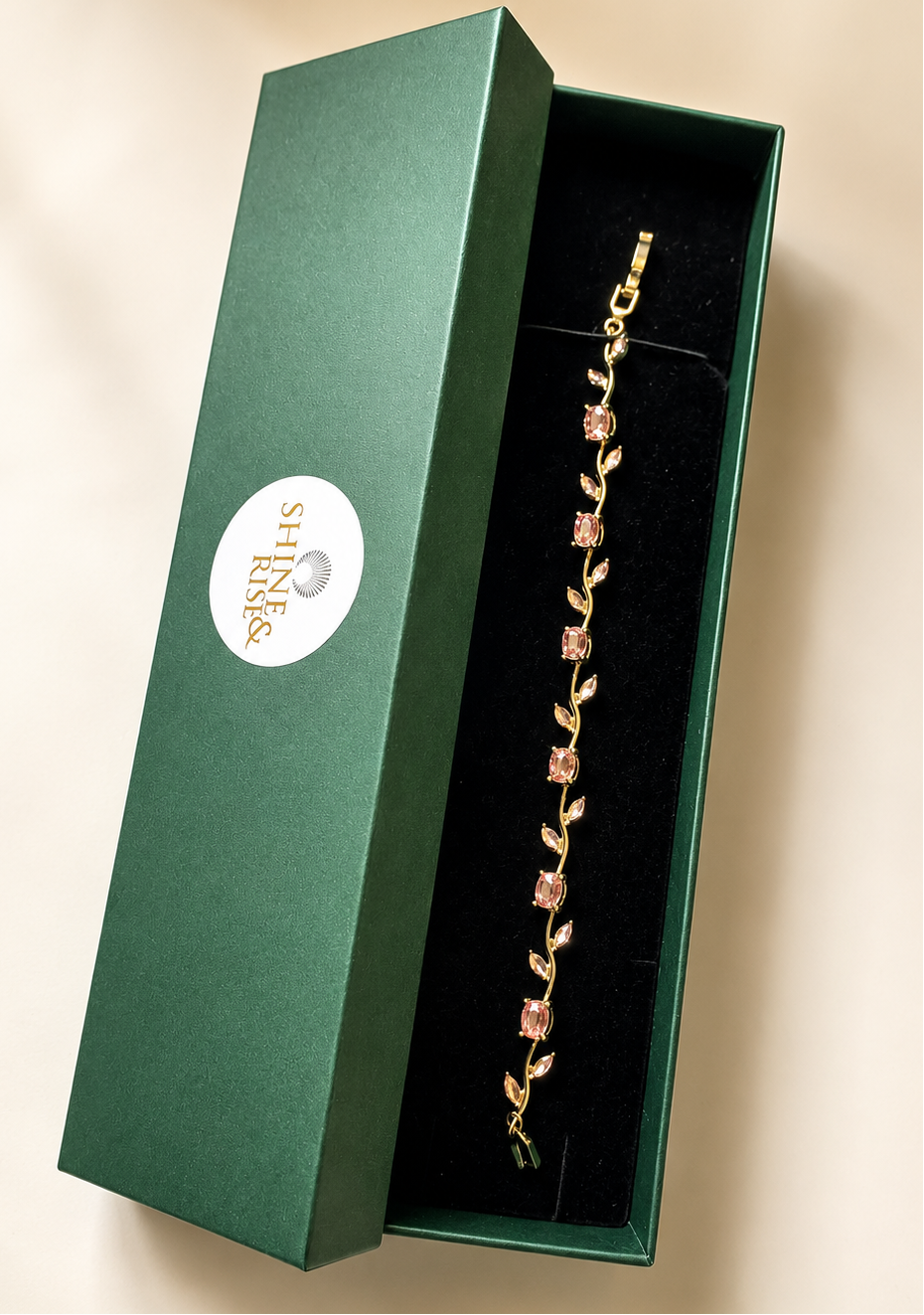 A gold bracelet with pink gemstone floral details displayed inside an open dark green jewelry box with a white circular logo.