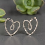 Heart-shaped earrings with sparkling stones on a gray surface with green plants in the background